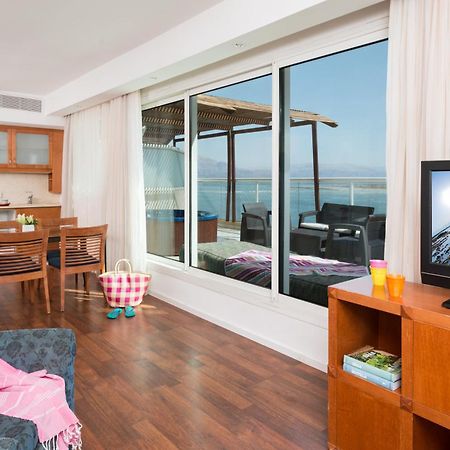 ليوناردو كلوب هوتيل البحر الميت - شامل كلياً نيفيه زوهار المظهر الخارجي الصورة The photo depicts a modern living space with a view. On one side, there is a comfortable seating area with a couch and a television cabinet. The TV is turned on, showing a scenic image. 

In the background, there is a dining area with a wooden table 