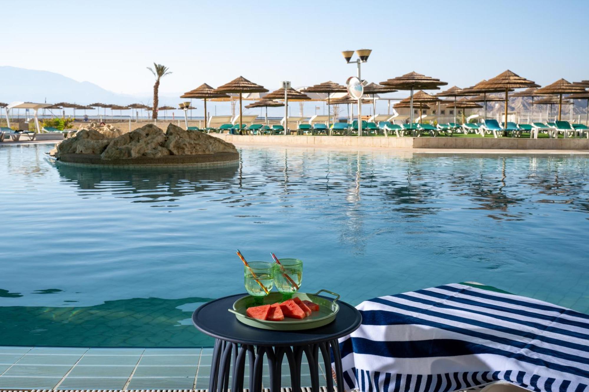 ليوناردو كلوب هوتيل البحر الميت - شامل كلياً نيفيه زوهار المظهر الخارجي الصورة The photo depicts a tranquil swimming pool area at a resort or hotel. In the foreground, there is a small round table with a plate containing slices of watermelon and two green drinks. The background features a calm pool with a small rock island in t