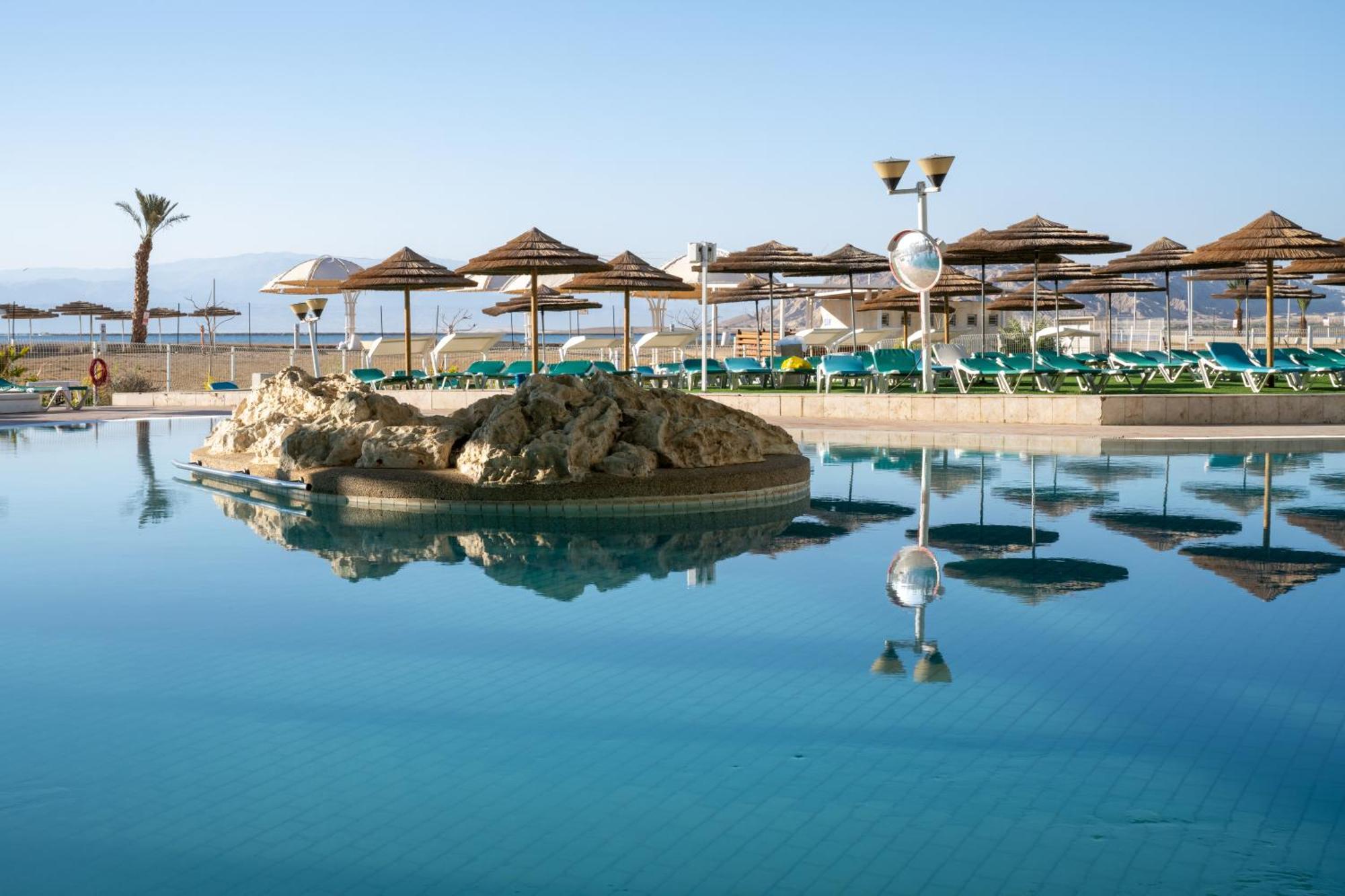 ليوناردو كلوب هوتيل البحر الميت - شامل كلياً نيفيه زوهار المظهر الخارجي الصورة The photo shows a tranquil swimming pool area. It features a large, reflective pool with a small rock formation in the center. Surrounding the pool are several sun loungers with green cushions and a series of thatched umbrellas providing shade. In th