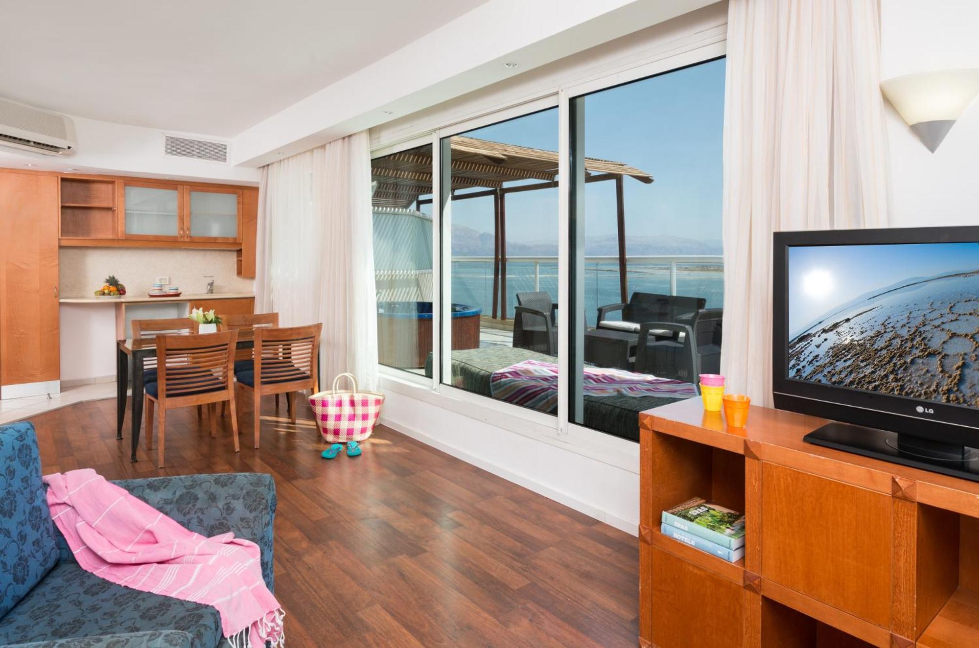 ليوناردو كلوب هوتيل البحر الميت - شامل كلياً نيفيه زوهار المظهر الخارجي الصورة The photo depicts a modern living space with a view. On one side, there is a comfortable seating area with a couch and a television cabinet. The TV is turned on, showing a scenic image. 

In the background, there is a dining area with a wooden table 